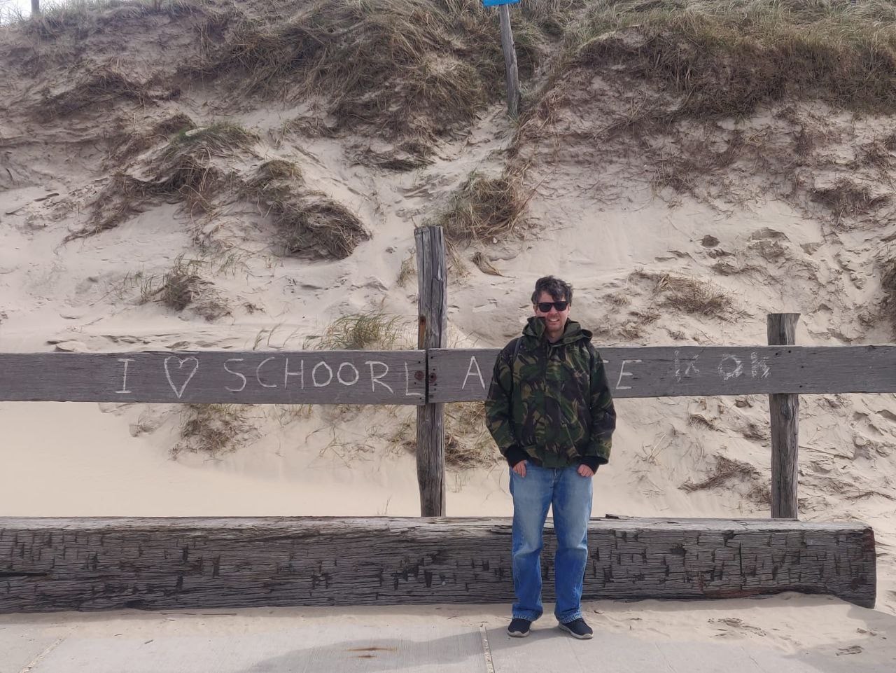 alex in camo jacket, on the beach at schoorl on sea, next to a sign saying I love Schoorl